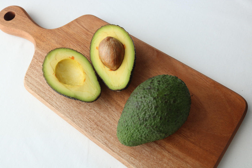 Avocado cut in half on a wooden cutting board with a whole avocado next to it.