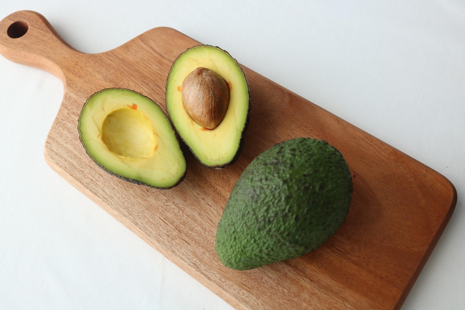 Avocado cut in half on a wooden cutting board with a whole avocado next to it.