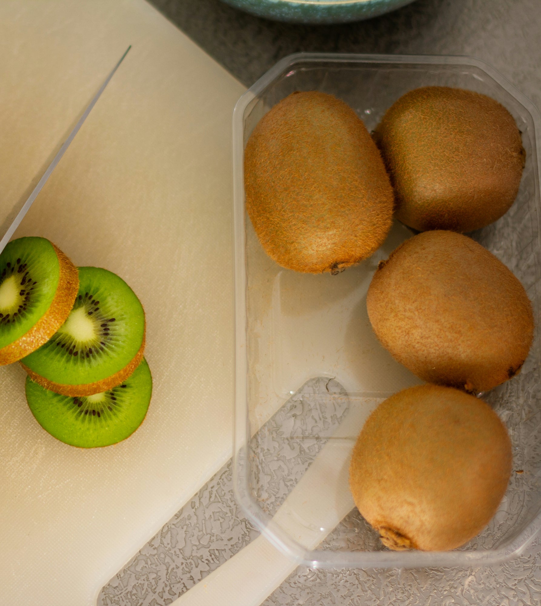 Three whole kiwi fruits in a clear plastic container next to sliced green kiwi on a white cutting board with a knife.