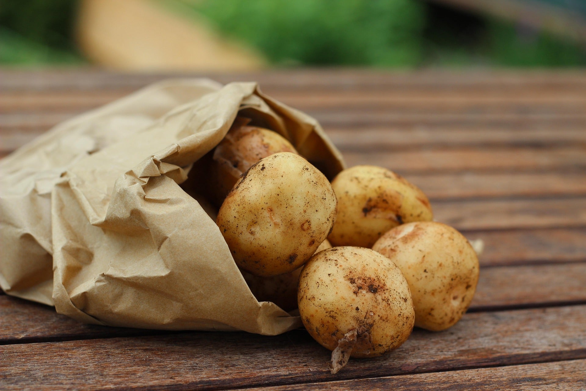 Fresh small potatoes spilling out of a brown paper bag onto a wooden table, with a softly blurred outdoor background.