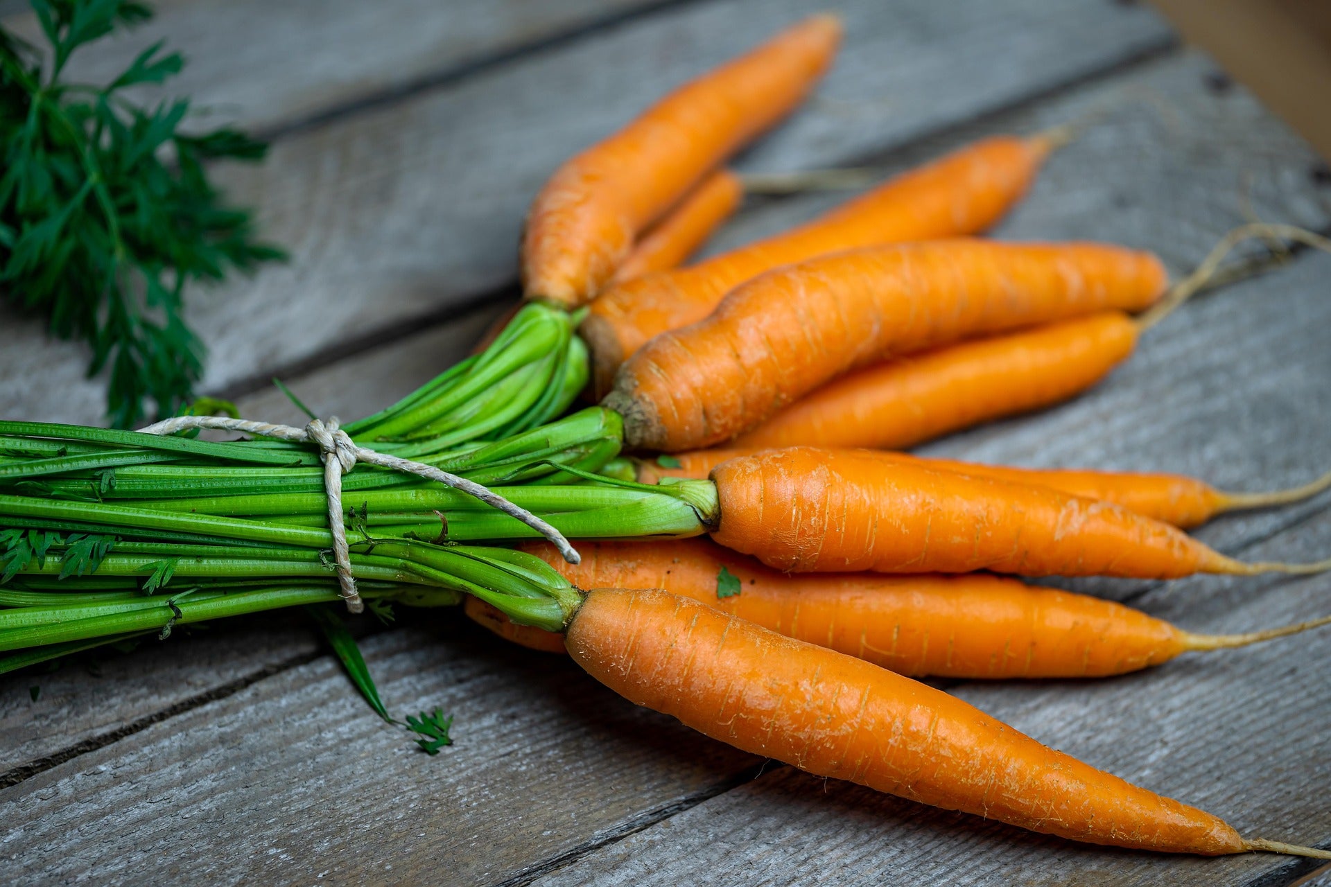 A bundle of fresh orange carrots with green tops tied together with string, placed on a rustic gray wooden surface.