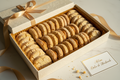 An open cream-colored gift box filled with rows of golden-brown dulce de mantecados (almond cookies), tied with a satin ribbon, on a light marble surface 