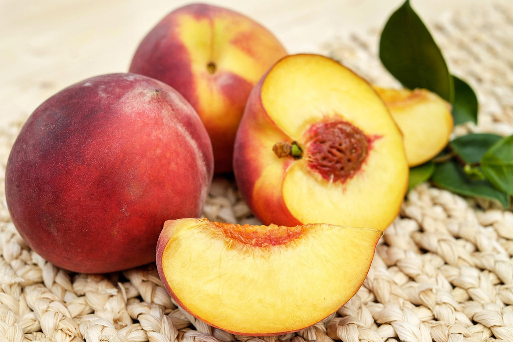 Fresh nectarines on a woven straw mat, including two whole nectarines, one sliced open showing the yellow-orange flesh and pit, and a couple of green leaves.