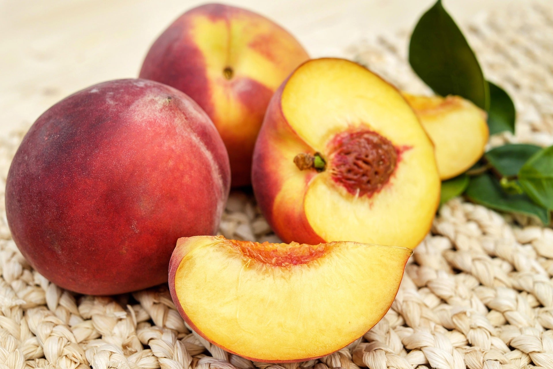 Fresh nectarines on a woven straw mat, including two whole nectarines, one sliced open showing the yellow-orange flesh and pit, and a couple of green leaves.