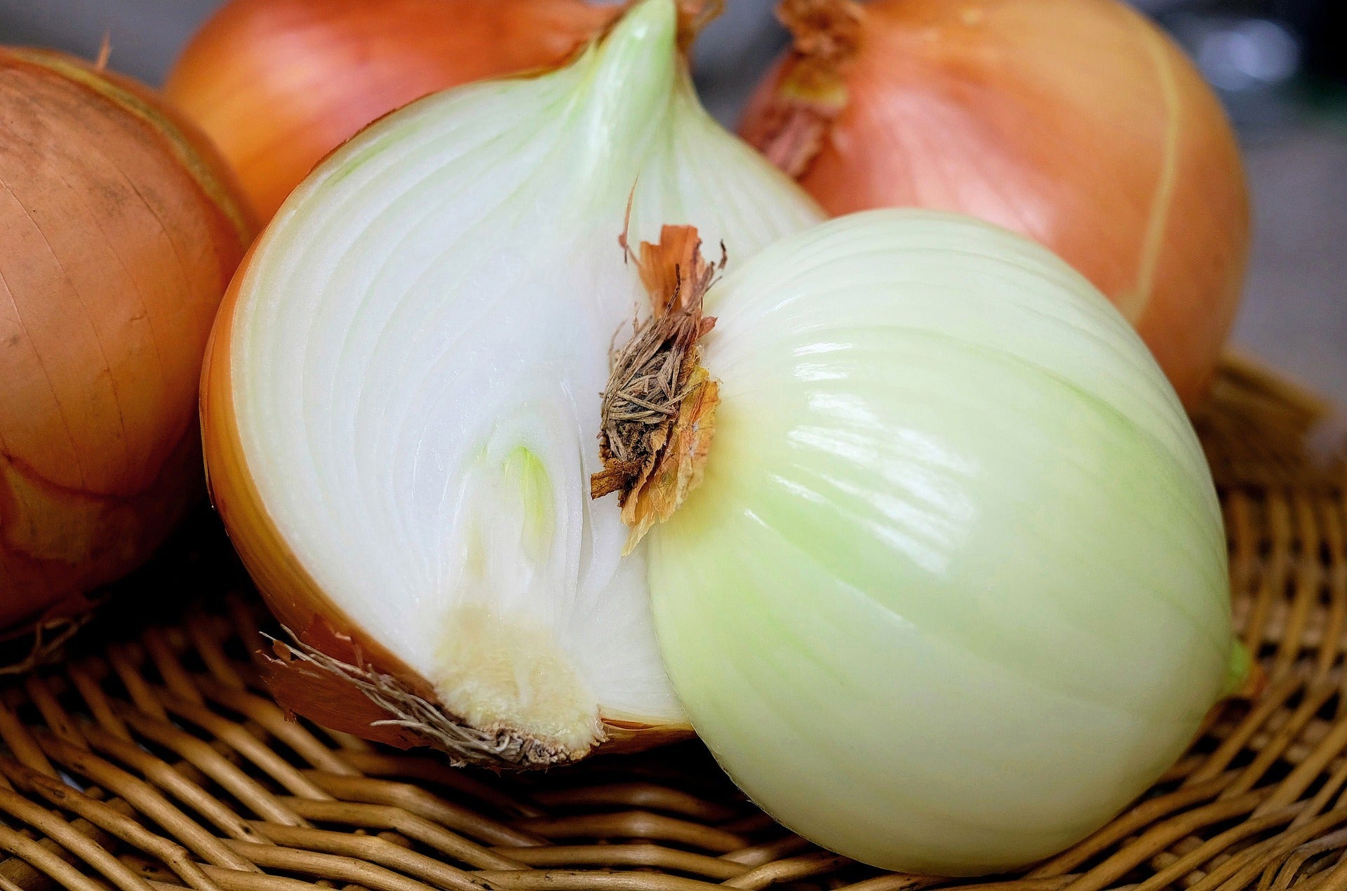 A close-up of fresh onions in a woven basket, including one halved onion showing its white layers and a whole pale green onion, with brown onion skins visible on the outer bulbs.