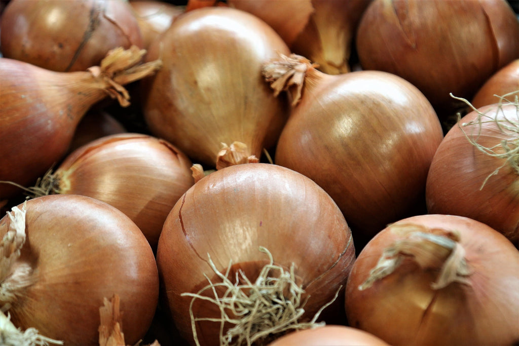 A close-up pile of whole yellow onions with golden-brown papery skins and a few dried green roots visible.