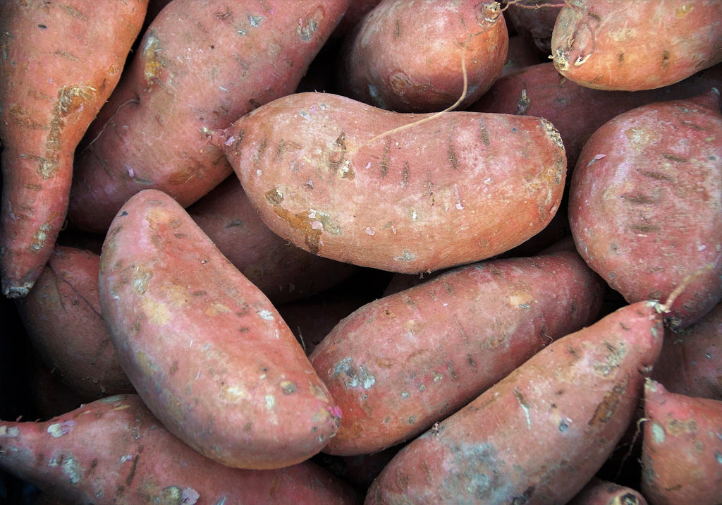 A close-up pile of sweet potatoes with rough, dusty reddish-brown skin filling the frame.